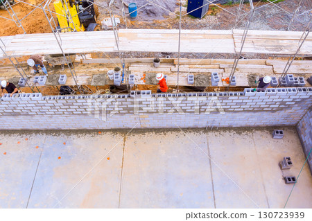 Several construction workers are laying concrete blocks to erect wall at busy construction site. 130723939
