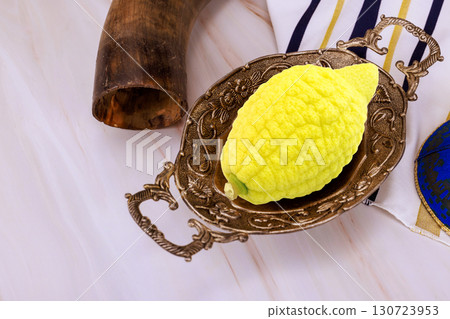 Yellow etrog in an ornate metal dish beside shofar, tallit gold stripes, decorative kippah on light marble surface. 130723953