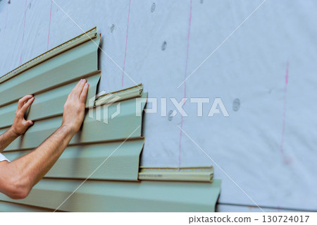 Workers are carefully aligning securing green PVC vinyl siding panels on exterior of home under renovation. Workers are carefully aligning securing green PVC vinyl siding panels on exterior of home under renovation. 130724017
