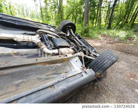 An overturned vehicle rests on its side in woods, damage from an accident its undercarriage amid greenery. 130724028
