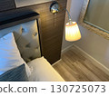 Cozy bedroom corner with a wall-mounted bedside lamp glowing warm light beside a tufted headboard, white pillows, and soft bedding, creating a relaxing and comfortable atmosphere for rest 130725073