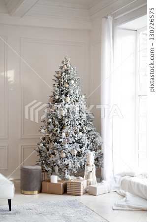 A beautifully decorated Christmas tree with white and gold ornaments and a snow effect, in a cozy and bright modern living room. 130726575