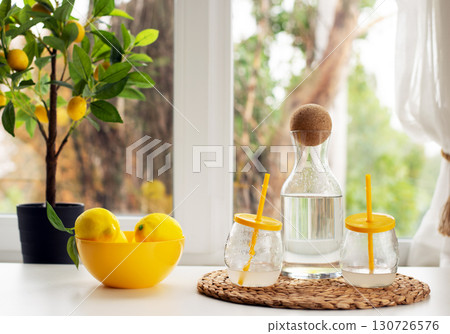 A healthy and fresh lifestyle scene with a carafe of water, lemons in a yellow bowl, and a small lemon tree on a bright kitchen counter. 130726576