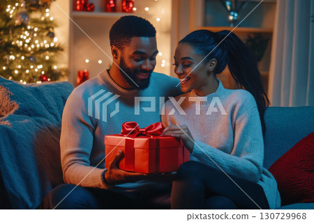 Happy diverse young couple exchanging a festive red gift on a cozy sofa during winter holidays, smiling with joy. 130729968