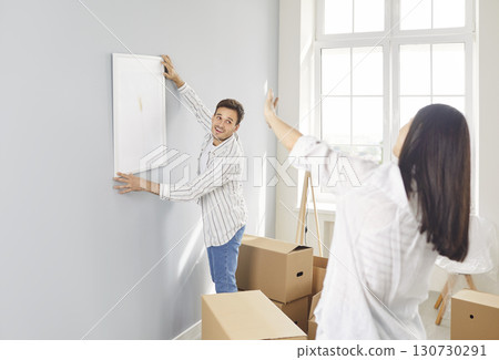 Happy young husband, wife laughing to enjoy hanging frame picture on wall, moving settling together 130730291