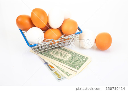 A wire shopping basket filled with white and brown eggs, placed beside rolled dollar bills A wire shopping basket filled with white and brown eggs, placed beside rolled dollar bills 130730414