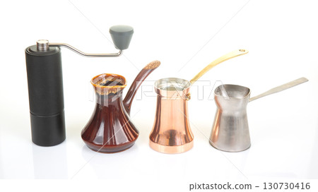 A black manual coffee grinder with a metallic handle stands beside a ceramic cezve A black manual coffee grinder with a metallic handle stands beside a ceramic cezve 130730416