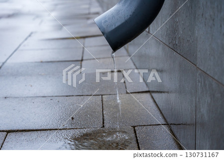 Rainwater drips from a dark downspout onto wet grey paving stones. Clear water flows from the building pipe, creating puddles on the sidewalk. 130731167