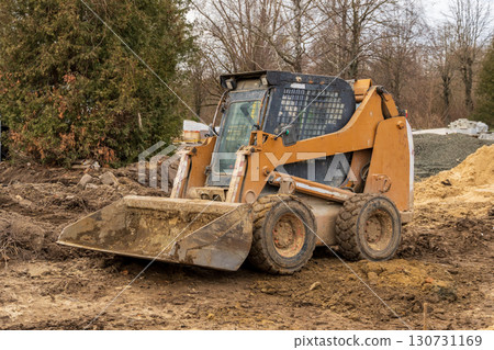 Orange skid steer loader parked on muddy ground. Heavy construction equipment with dirty bucket on a site with dirt piles and trees. Orange skid steer loader parked on muddy ground. Heavy construction equipment with dirty bucket on a site with dirt piles and trees. 130731169
