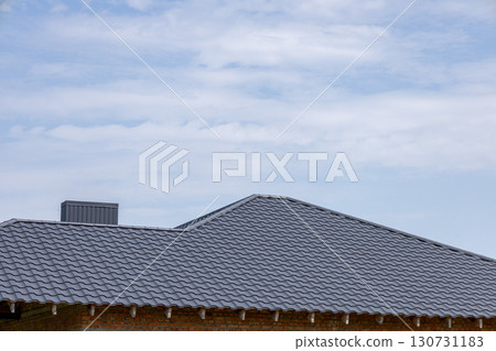 Modern house roof with dark grey tiles, featuring a chimney and brick wall. New residential building construction under a cloudy blue sky. 130731183