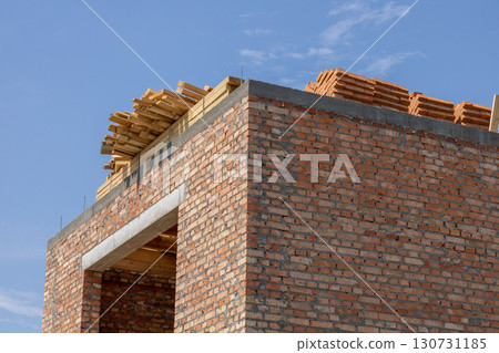 Developing house construction project showing unfinished red brick walls and wooden roof framing. Stacked building materials under a clear blue sky. Developing house construction project showing unfinished red brick walls and wooden roof framing. Stacked building materials under a clear blue sky. 130731185