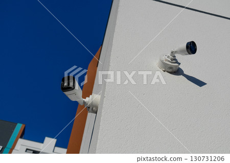 Two white security cameras mounted on textured building wall under a clear blue sky. Modern surveillance technology for urban security. Two white security cameras mounted on textured building wall under a clear blue sky. Modern surveillance technology for urban security. 130731206