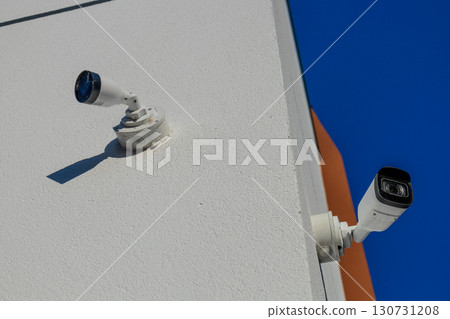White security camera and outdoor light fixture mounted on a textured building wall. Clear blue sky, bright sun, long shadows. White security camera and outdoor light fixture mounted on a textured building wall. Clear blue sky, bright sun, long shadows. 130731208
