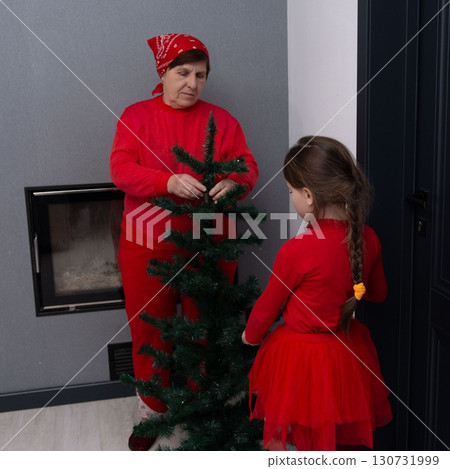 Grandmother and granddaughter decorating a small Christmas tree together, wearing matching red outfits. Grandmother and granddaughter decorating a small Christmas tree together, wearing matching red outfits. 130731999