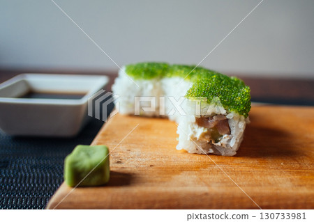 A sushi roll with green seaweed on top sits on a wooden cutting board 130733981