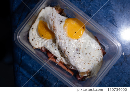 Simple meal of two fried eggs placed over grilled sausage, served in a clear plastic container. 130734000