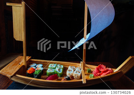 A wooden boat with a sail is filled with various sushi rolls 130734026