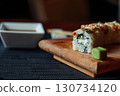 A sushi roll sits on a wooden cutting board with a small green leaf on top 130734120