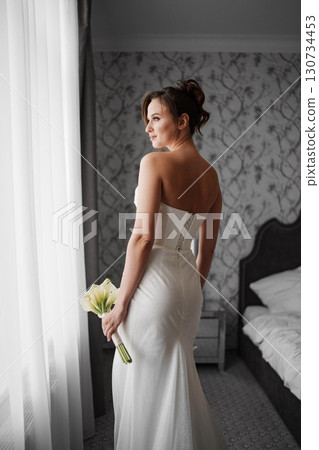 bride in a tight-fitting dress with a bouquet calla by windows in a hotel room. 130734453