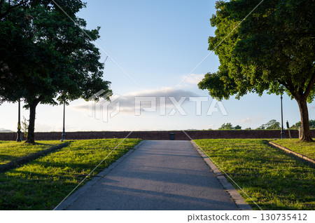 Evening walk along the historic city walls of Lucca, Tuscany, Italy. Lined with trees, the path glows in the warm light of the setting sun, offering a tranquil atmosphere. Evening walk along the historic city walls of Lucca, Tuscany, Italy. Lined with trees, the path glows in the warm light of the setting sun, offering a tranquil atmosphere. 130735412
