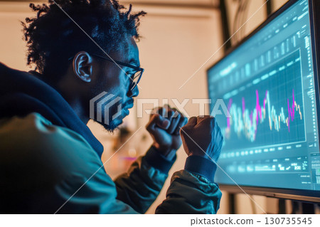 Young Black man with glasses stares intently at computer screen displaying glowing financial charts. He clenches fists, feeling success and determination. Young Black man with glasses stares intently at computer screen displaying glowing financial charts. He clenches fists, feeling success and determination. 130735545