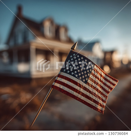 A small American flag gently waves on its pole, standing before a blurred house in warm outdoor light. 130736084