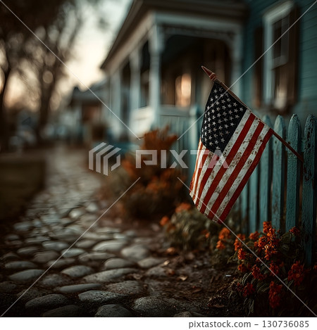 A rustic American flag hangs on a weathered fence with a quaint house and stone path in the background. 130736085