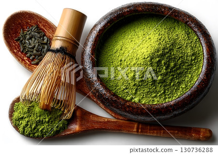 Bright green matcha powder fills a dark wooden bowl, alongside a bamboo whisk and tea leaves on wooden spoons. 130736288