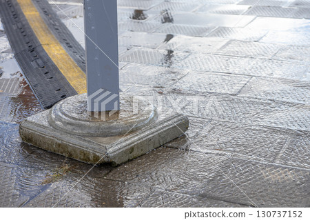 Water flows across grey textured urban paving, highlighting a concrete pole base and a black-yellow cable protector, depicting city drainage 130737152