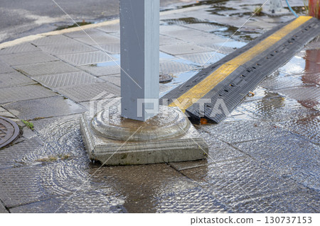 Wet city sidewalk pavement with water pooling around a grey utility pole base and a black cable protector, showing urban drainage challenges 130737153