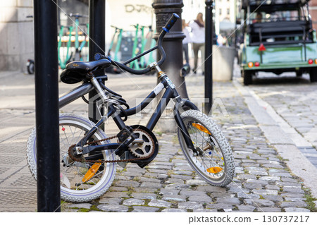 Black bicycle with small wheels securely locked to a metal pole, parked on a historic cobblestone street, portraying urban transport and city life Black bicycle with small wheels securely locked to a metal pole, parked on a historic cobblestone street, portraying urban transport and city life 130737217
