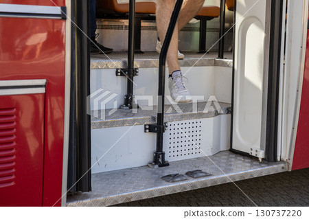 A passenger's bare leg in white sneakers steps down the metal stairs of a red public bus, depicting urban mobility, daily commuting, and accessibility in travel A passenger's bare leg in white sneakers steps down the metal stairs of a red public bus, depicting urban mobility, daily commuting, and accessibility in travel 130737220