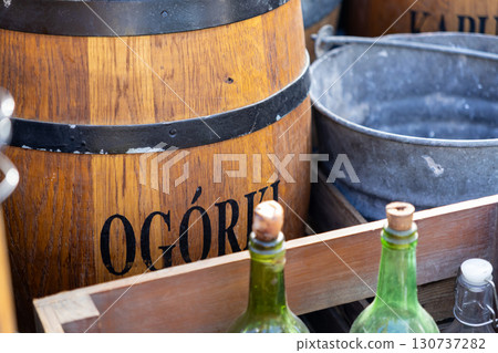 Close-up of a rustic wooden barrel with OGORKI text, vintage green bottles, and metal bucket, reflecting traditional food market culture 130737282