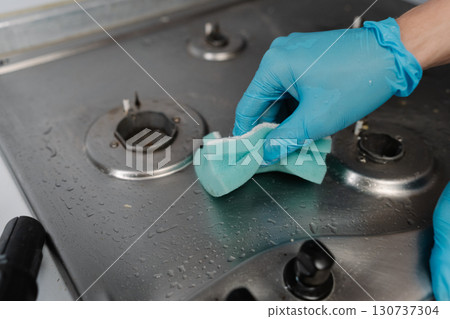 Professional housekeeper wearing blue gloves scrubbing dirty gas stove with sponge, applying cleaning solution for sparkling kitchen surfaces, close-up. Concept of cleanliness and housekeeping. Professional housekeeper wearing blue gloves scrubbing dirty gas stove with sponge, applying cleaning solution for sparkling kitchen surfaces, close-up. Concept of cleanliness and housekeeping. 130737304