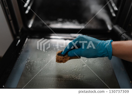 Professional cleaning worker wearing blue glove using sponge for cleaning glass door of gas oven, performing meticulous kitchen appliance maintenance and sanitization, close-up. Professional cleaning worker wearing blue glove using sponge for cleaning glass door of gas oven, performing meticulous kitchen appliance maintenance and sanitization, close-up. 130737309