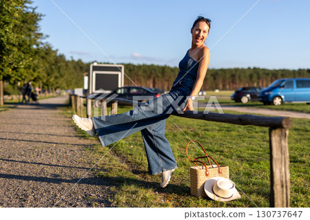 Young adult beautiful woman wear stylish jeans one-shoulder denim jumpsuit have fun sit outdoors warm sunny day holding straw bag hat hand. Female person carefree smiles enjoying countryside nature 130737647