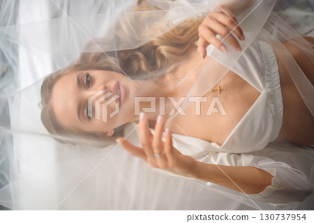 Beautiful blonde bride in white silk pajamas in bed under veil. 130737954
