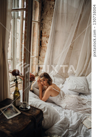 a young blonde woman in a white dress on white sheets by the window 130738004