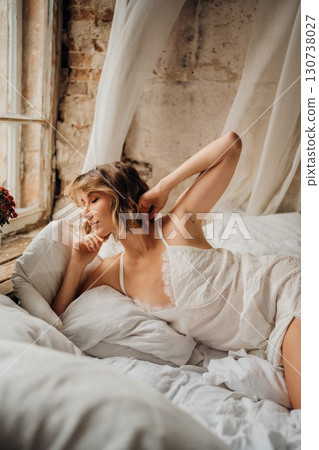 a blonde woman in a white dress on white sheets by the window 130738027