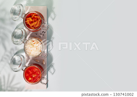 Probiotic food. Pickled or fermented vegetables. Lecho, sauerkraut, ajvar in glass jars on a white board on a blue background with shadows and copy space. Home food preserving or canning. Close up Probiotic food. Pickled or fermented vegetables. Lecho, sauerkraut, ajvar in glass jars on a white board on a blue background with shadows and copy space. Home food preserving or canning. Close up 130741002