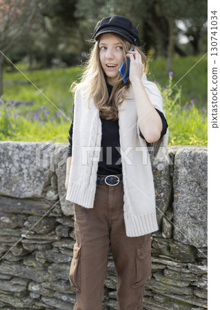 Teenage girl talking on smartphone wearing newsboy cap and knitted vest Teenage girl talking on smartphone wearing newsboy cap and knitted vest 130741034