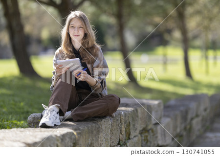 Young woman writing in notebook sitting on stone wall in park 130741055