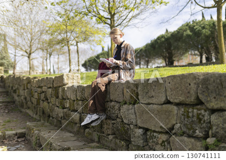 Young student taking notes while sitting on stone wall in park 130741111