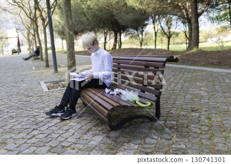 Young student studying on park bench with folded umbrella Young student studying on park bench with folded umbrella 130741301