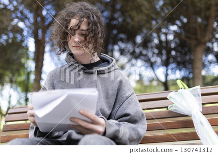 Young man reading documents on park bench with folded umbrella 130741312