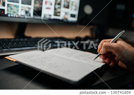 Closeup of hand writing notes in notebook at desk with computer. Concept of planning, studying, working and productivity. 130741804