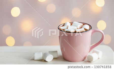 Pink mug filled with hot chocolate and marshmallows on light wooden table with festive glowing bokeh lights in soft background 130741858