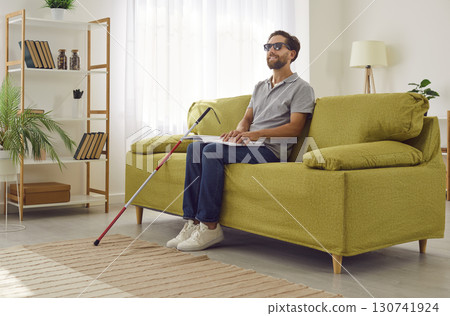 Blind man in dark eyeglasses sitting on sofa at home and reading tactile braille book. Blind man in dark eyeglasses sitting on sofa at home and reading tactile braille book. 130741924