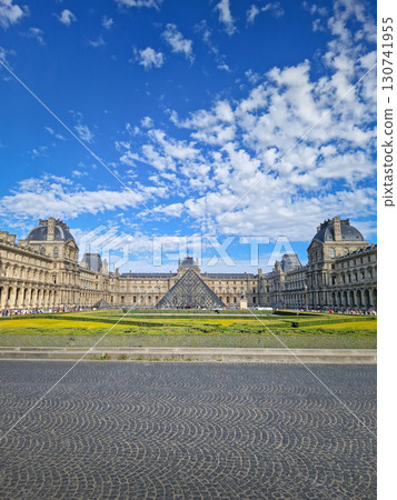 A grand panoramic view of the Louvre Palace with its iconic glass pyramid in the center under a magnificent blue sky with fluffy white clouds in Paris, France 130741955