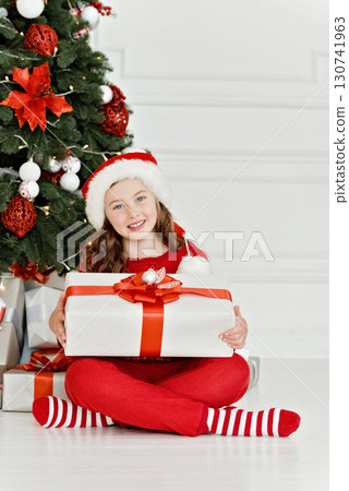 Smiling girl in Santa hat and red pajamas sitting by decorated Christmas tree holding a large white gift box with red ribbon Smiling girl in Santa hat and red pajamas sitting by decorated Christmas tree holding a large white gift box with red ribbon 130741963